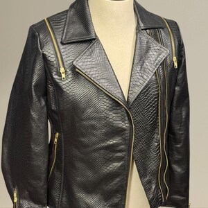 K&ouml;nnte beinhalten: Schwarze Kunstlederjacke mit Schlangenlederstruktur. Die Jacke hat einen Reverskragen, einen asymmetrischen Rei&szlig;verschluss und mehrere goldfarbene Rei&szlig;verschl&uuml;sse. Die Jacke wird auf einer Schaufensterpuppe pr&auml;sentiert.