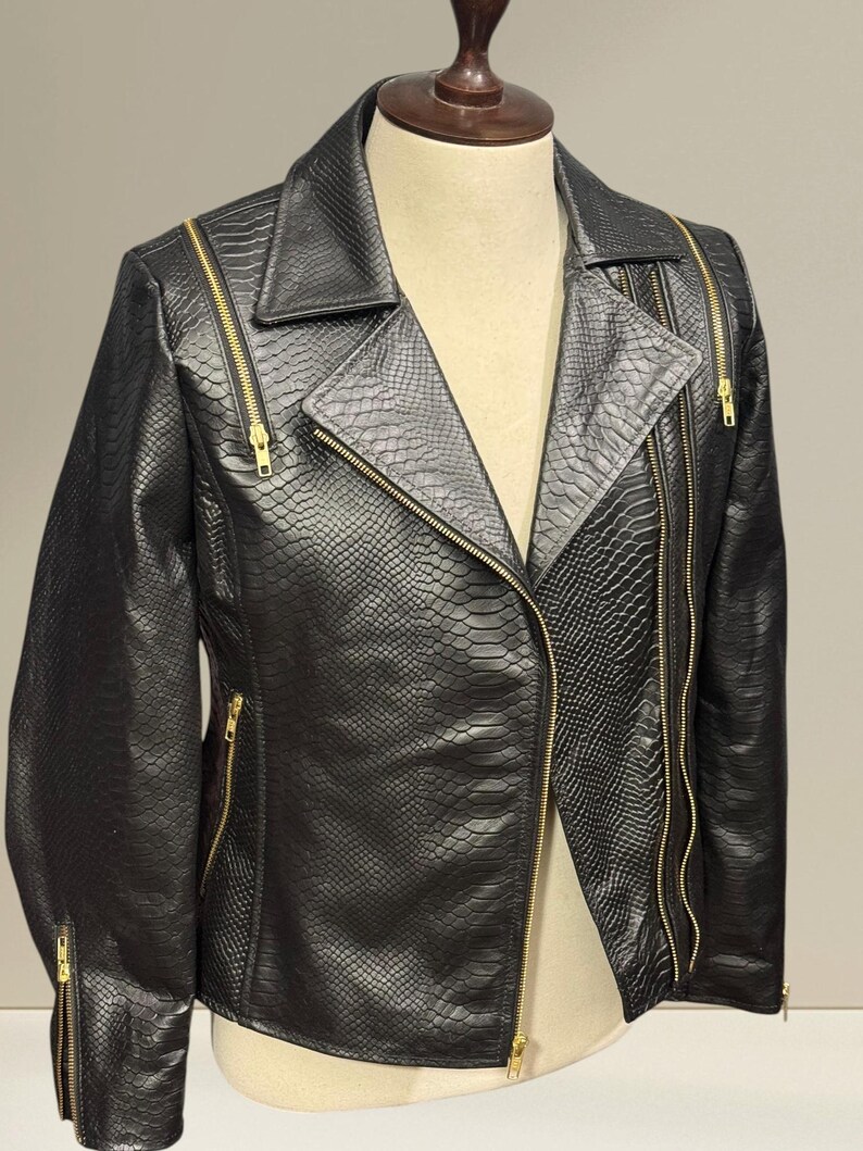 K&ouml;nnte beinhalten: Schwarze Kunstlederjacke mit Schlangenlederstruktur. Die Jacke hat einen Reverskragen, einen asymmetrischen Rei&szlig;verschluss und mehrere goldfarbene Rei&szlig;verschl&uuml;sse. Die Jacke wird auf einer Schaufensterpuppe pr&auml;sentiert.