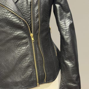 K&ouml;nnte beinhalten: Schwarze Lederjacke mit Schlangenleder-Textur und goldenen Rei&szlig;verschl&uuml;ssen. Die Jacke hat ein klassisches asymmetrisches Design mit einem Reverskragen und mehreren Rei&szlig;verschlusstaschen. Die &Auml;rmel sind lang und eng anliegend.