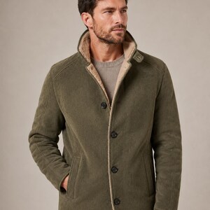 May include: An olive green jacket with a high collar and a beige faux-fur lining. The jacket features a button-front closure with four dark buttons, long sleeves, and two side pockets. It is worn over a light brown crew-neck sweater.