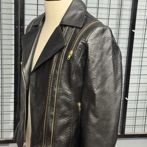 May include: Black faux leather jacket with a snakeskin texture. The jacket features a notched collar, multiple gold-toned zippers, and a classic biker jacket design. The jacket is displayed on a mannequin.