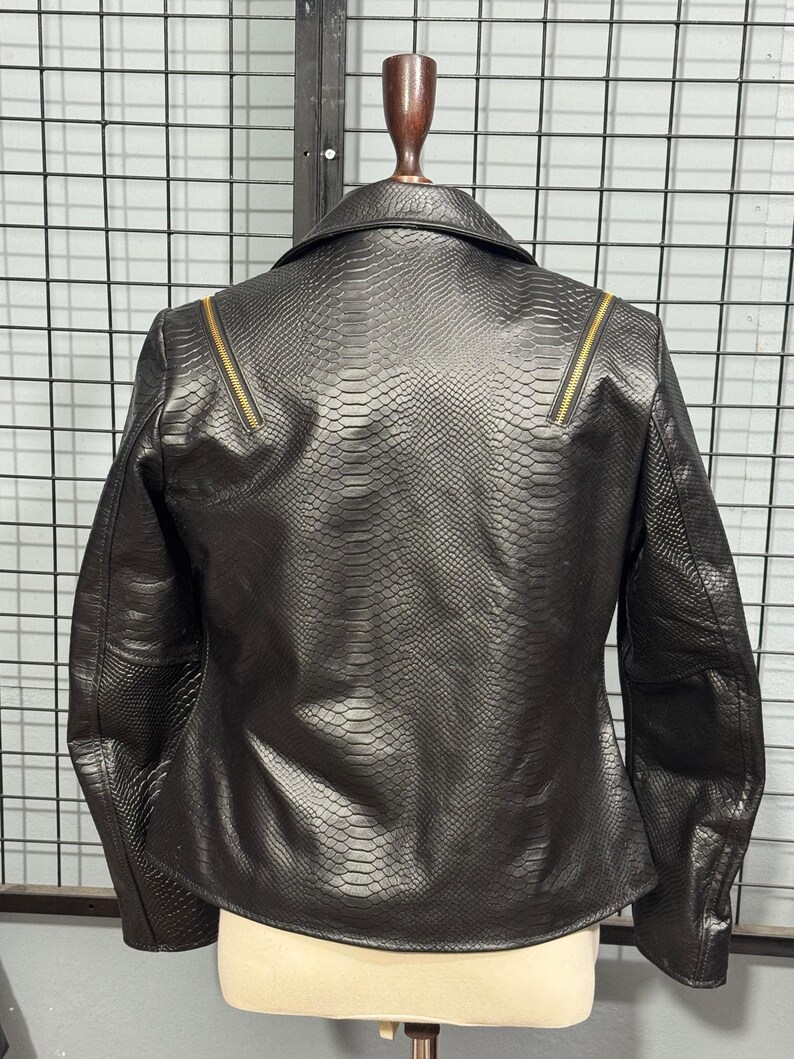 May include: Black faux leather jacket with a snakeskin texture. The jacket features a classic collar, long sleeves, and gold-toned zippers on the shoulders. The back view showcases the jacket's tailored fit and detailed design.
