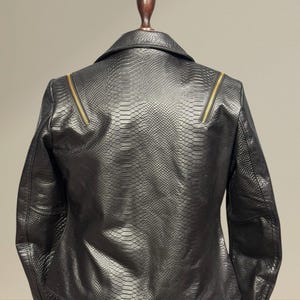 K&ouml;nnte beinhalten: Schwarze Kunstlederjacke mit Schlangenlederstruktur. Die Jacke hat einen klassischen Kragen, lange &Auml;rmel und goldfarbene Rei&szlig;verschl&uuml;sse an den Schultern. Die R&uuml;ckansicht der Jacke wird auf einer Schaufensterpuppe gezeigt.