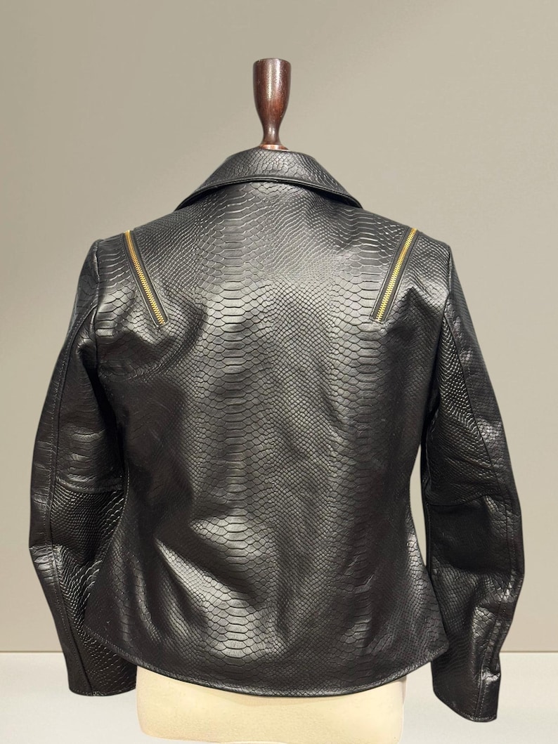 K&ouml;nnte beinhalten: Schwarze Kunstlederjacke mit Schlangenlederstruktur. Die Jacke hat einen klassischen Kragen, lange &Auml;rmel und goldfarbene Rei&szlig;verschl&uuml;sse an den Schultern. Die R&uuml;ckansicht der Jacke wird auf einer Schaufensterpuppe gezeigt.