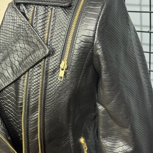May include: Black faux leather jacket with a snakeskin texture. The jacket features a classic asymmetrical design with a notched lapel, multiple zippered pockets, and gold-toned zippers. The jacket is a stylish outerwear piece.