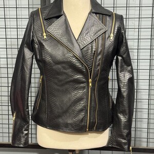 May include: Black faux leather jacket with a snakeskin texture and gold zippers. The jacket features an asymmetrical zipper closure, a notched collar, and multiple zippered pockets. The sleeves have zippers at the cuffs. The jacket is displayed on a mannequin.