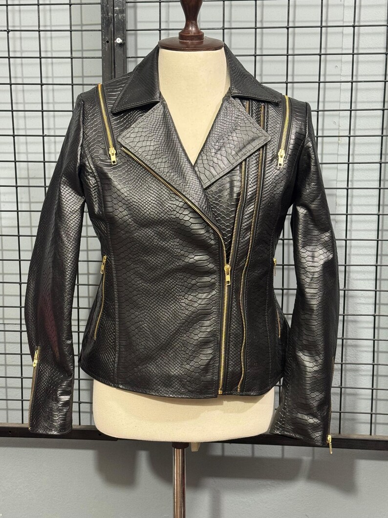 May include: Black faux leather jacket with a snakeskin texture and gold zippers. The jacket features an asymmetrical zipper closure, a notched collar, and multiple zippered pockets. The sleeves have zippers at the cuffs. The jacket is displayed on a mannequin.