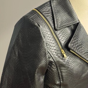 K&ouml;nnte beinhalten: Schwarze Kunstlederjacke mit Schlangenlederstruktur. Die Jacke hat ein asymmetrisches Rei&szlig;verschlussdesign mit goldfarbenen Rei&szlig;verschl&uuml;ssen. Der Kragen ist ein Reverskragen. Die Jacke wird auf einer Schaufensterpuppe pr&auml;sentiert.
