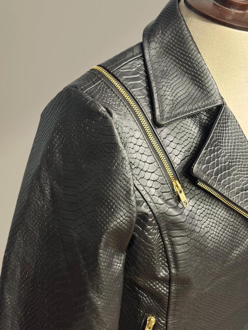 K&ouml;nnte beinhalten: Schwarze Kunstlederjacke mit Schlangenlederstruktur. Die Jacke hat ein asymmetrisches Rei&szlig;verschlussdesign mit goldfarbenen Rei&szlig;verschl&uuml;ssen. Der Kragen ist ein Reverskragen. Die Jacke wird auf einer Schaufensterpuppe pr&auml;sentiert.