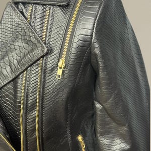 K&ouml;nnte beinhalten: Nahaufnahme einer schwarzen Lederjacke mit Schlangenlederstruktur. Die Jacke hat einen Reverskragen, mehrere goldfarbene Rei&szlig;verschl&uuml;sse und eine taillierte Passform. Das Gesamtdesign ist elegant und kantig.