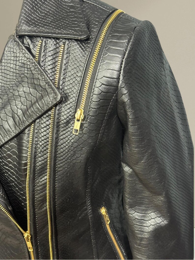 K&ouml;nnte beinhalten: Nahaufnahme einer schwarzen Lederjacke mit Schlangenlederstruktur. Die Jacke hat einen Reverskragen, mehrere goldfarbene Rei&szlig;verschl&uuml;sse und eine taillierte Passform. Das Gesamtdesign ist elegant und kantig.