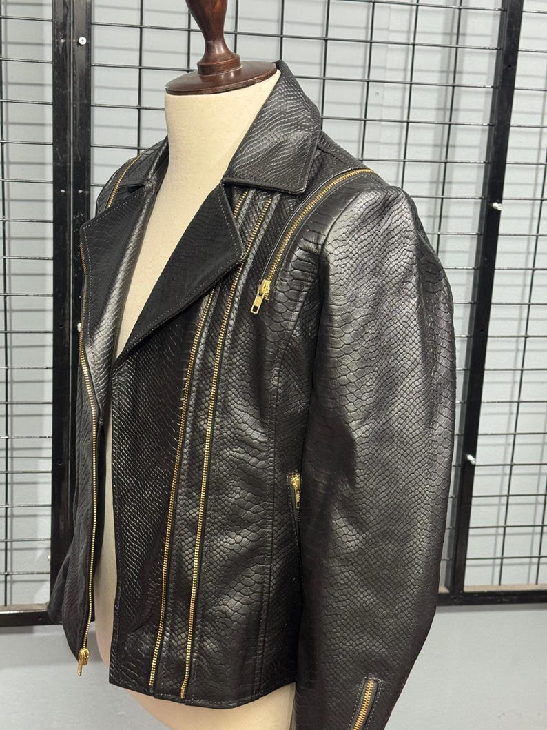 May include: Black faux leather jacket with a snakeskin texture. The jacket features a notched collar, multiple gold zippers, and a classic biker style. The jacket is displayed on a mannequin.