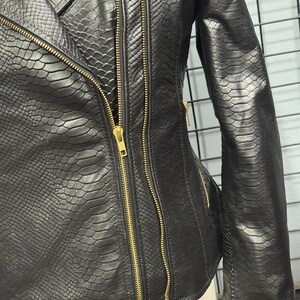 May include: Black faux leather jacket with a snakeskin texture. The jacket features a diagonal zipper closure, multiple gold zippers, and a tailored fit. The sleeves are long, and the jacket has a classic biker style.