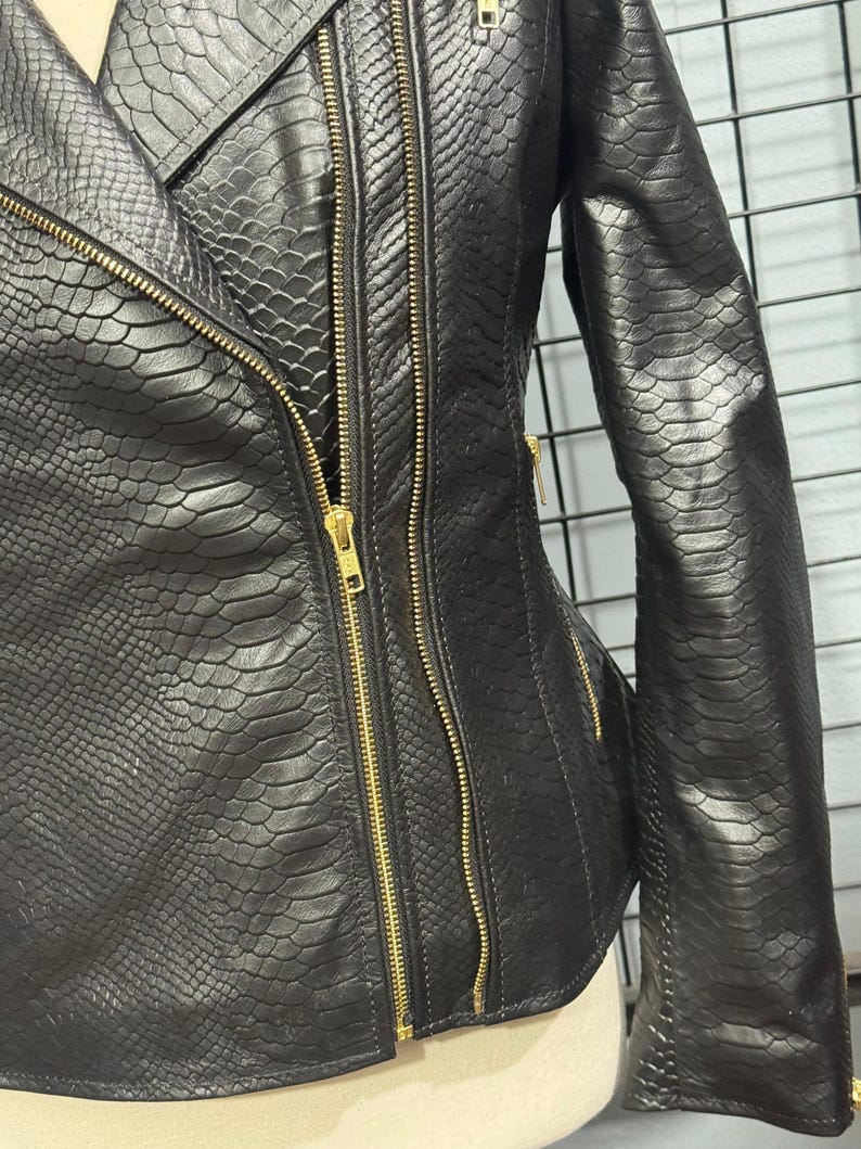 May include: Black faux leather jacket with a snakeskin texture. The jacket features a diagonal zipper closure, multiple gold zippers, and a tailored fit. The sleeves are long, and the jacket has a classic biker style.