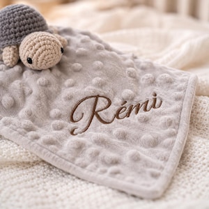Peut inclure: Une couverture de sécurité en peluche gris clair avec un motif à pois texturé et une tête de tortue crochetée attachée au coin. La couverture présente le prénom Rémi brodé en lettres cursives marron au centre de la surface en tissu doux.