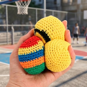 Può includere: Una mano tiene tre palline da giocoleria all'uncinetto. Una è gialla con una striscia nera, un'altra è a righe rosse, arancioni, blu e verdi, e la terza è giallo pieno.