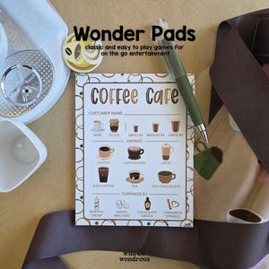 Peut inclure: Une carte de jeu "Coffee Cafe" avec un menu de boissons et de garnitures de café, un stylo vert et le texte "Wonder Pads" en haut. La carte est sur une surface brun clair avec un appareil blanc et un ruban marron.