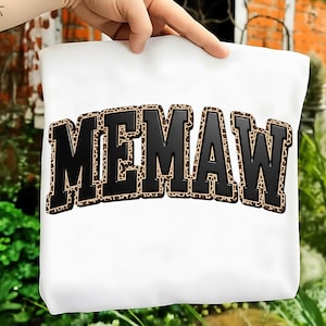 May include: White tote bag with the word "MEMAW" in large, black, block letters. The letters are outlined with a leopard print pattern. The bag is held up by a hand.