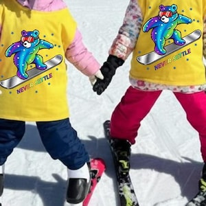 Può includere: Due bambini piccoli con magliette gialle con un orso colorato su uno snowboard, caschi neri e attrezzatura da sci. Sono in piedi su una pista innevata, tenendosi per mano, con edifici sullo sfondo.