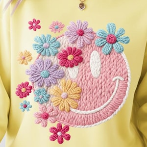 Puede incluir: Una sudadera amarilla clara con una gran cara sonriente rosa bordada adornada con flores coloridas. Las flores incluyen tonos de rosa, azul, morado y amarillo. La sudadera tiene mangas largas y cuello redondo.