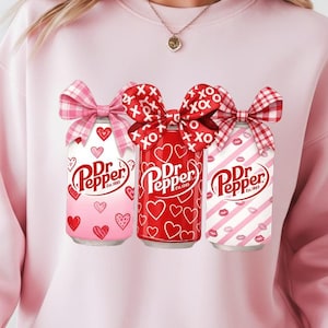May include: Pink sweatshirt featuring three Dr. Pepper cans with Valentine's Day designs. The cans have bows and are decorated with hearts, XOXO, and lip prints. The sweatshirt is a light pink color.