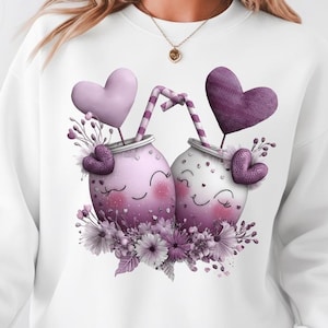 Pode incluir: Camisola branca com uma ilustração lúdica de duas canecas sorridentes com coberturas em forma de coração e detalhes florais. As canecas são em tons de roxo e rosa, conectadas por canudos listrados.