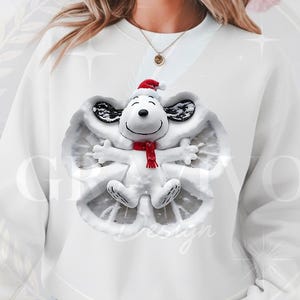 May include: White sweatshirt featuring a Snoopy angel design. Snoopy is wearing a Santa hat and a red scarf. The design is surrounded by a snow angel shape. The sweatshirt is a festive holiday item.