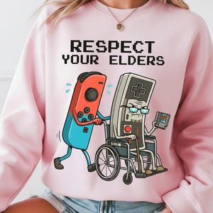 Peut inclure: Un sweat-shirt rose clair avec le texte "RESPECT YOUR ELDERS" au-dessus d'un dessin animé. Le dessin animé représente une manette de jeu rouge et bleue poussant une manette grise en fauteuil roulant. La manette grise porte des lunettes et tient une cartouche de jeu.