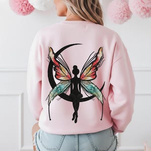 May include: A light pink sweatshirt featuring a black silhouette of a fairy sitting on a crescent moon. The fairy has colorful wings with red, yellow, and blue accents. The sweatshirt is a casual style.