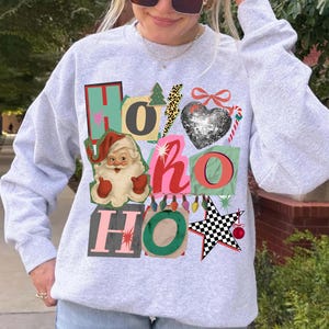 Könnte beinhalten: Hellgraues Sweatshirt mit festlichem "Ho Ho Ho"-Design. Das Design umfasst eine Weihnachtsmann-Grafik, ein paillettenbesetztes Herz, einen karierten Stern und andere weihnachtliche Elemente in verschiedenen Farben.