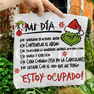 Puede incluir: Bolsa de mano blanca con el texto "MI DÍA" y un gráfico del Grinch con un gorro de Papá Noel. La bolsa presenta un horario de actividades en español, incluyendo "ESTOY OCUPADO!" y una guirnalda de luces.
