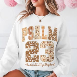 Puede incluir: Sudadera blanca con el texto "PSALM 23" en una mezcla de estampados florales, de vetas de madera y cuadros vichy. El número "23" tiene un estampado floral. El texto "The Lord Is My Shepherd" está debajo.
