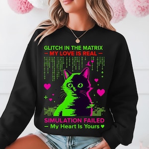 Puede incluir: Sudadera negra con un gráfico de un gato dividido en verde neón y morado. El texto dice "GLITCH IN THE MATRIX - MY LOVE IS REAL" y "SIMULATION FAILED - My Heart Is Yours". Corazones rosas y código binario completan el diseño.