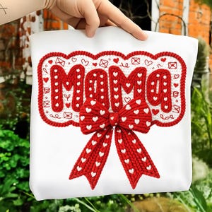 May include: White tote bag with the word "MAMA" in red, heart-filled letters. A large red bow with white hearts is at the bottom. The letters and bow are made of a textured material, possibly knitted or crocheted.