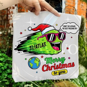 May include: A light gray tote bag featuring a cartoon pickle wearing sunglasses and a Santa hat. The pickle has the text "31/ATLAS" and the speech bubble says "DON'T BE AFRAID, I'M JUST PASSING BY!". The words "Merry Christmas to you" are also on the bag.