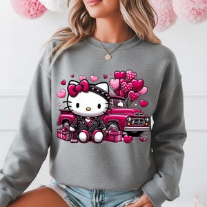 Puede incluir: Sudadera gris con un dise&ntilde;o de Hello Kitty. Lleva una chaqueta de cuero negra y un lazo rosa, sentada frente a una camioneta rosa con globos en forma de coraz&oacute;n y cajas de regalo. Ideal para el D&iacute;a de San Valent&iacute;n, el dise&ntilde;o es encantador.