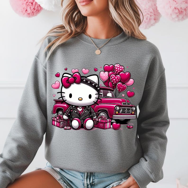 Puede incluir: Sudadera gris con un dise&ntilde;o de Hello Kitty. Lleva una chaqueta de cuero negra y un lazo rosa, sentada frente a una camioneta rosa con globos en forma de coraz&oacute;n y cajas de regalo. Ideal para el D&iacute;a de San Valent&iacute;n, el dise&ntilde;o es encantador.