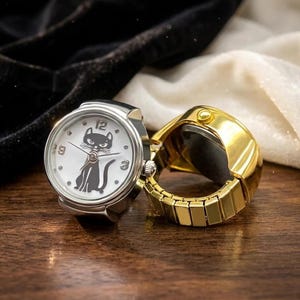 May include: Two ring watches. One is silver-toned with a white face featuring a black cat illustration and black numbers. The other is gold-toned. Both have a round watch face and a band that expands to fit the finger.