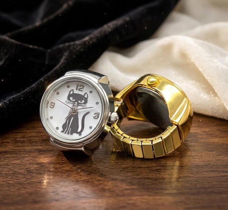 May include: Two ring watches. One is silver-toned with a white face featuring a black cat illustration and black numbers. The other is gold-toned. Both have a round watch face and a band that expands to fit the finger.