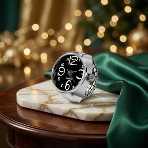 May include: A silver ring watch with a black face and white numerals. The watch is set on a white marble surface with gold veining. The background features a green satin fabric and blurred golden lights.