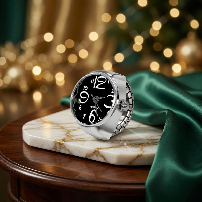 May include: A silver ring watch with a black face and white numerals. The watch is set on a white marble surface with gold veining. The background features a green satin fabric and blurred golden lights.