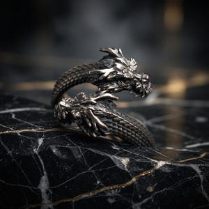 May include: A silver-toned dragon ring with intricate detailing. The ring features two dragon heads facing opposite directions, with textured scales and detailed facial features. The ring is displayed on a black and white marble surface.