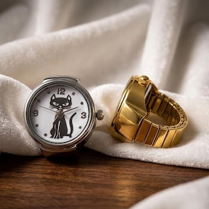 May include: A silver ring watch with a white face featuring a black cat silhouette. The watch has black hands and the numbers 3, 9, and 12. A gold-colored ring watch is next to it, with a band made of rectangular links.