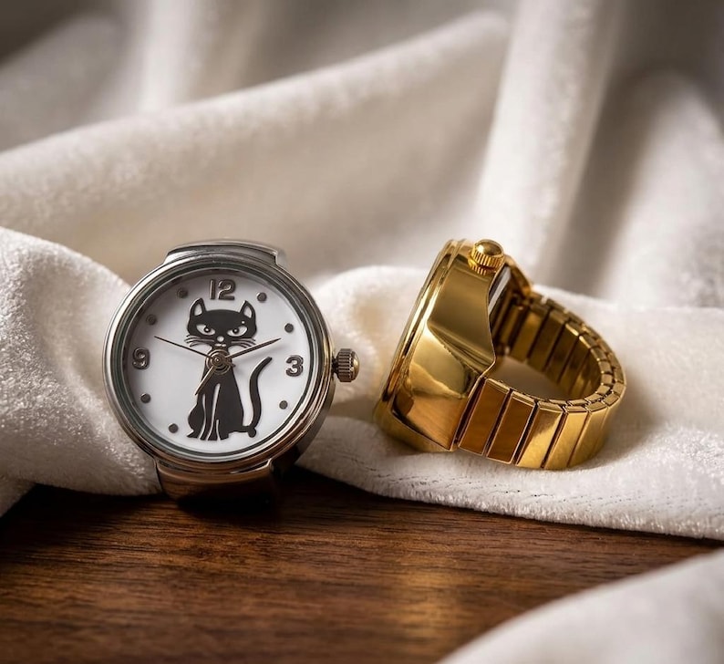 May include: A silver ring watch with a white face featuring a black cat silhouette. The watch has black hands and the numbers 3, 9, and 12. A gold-colored ring watch is next to it, with a band made of rectangular links.