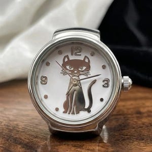 May include: A silver-toned ring watch with a white face. The watch features a cat silhouette at the center, with the numbers 3, 9, and 12 visible. The watch has a small crown on the side and is set on a wooden surface.