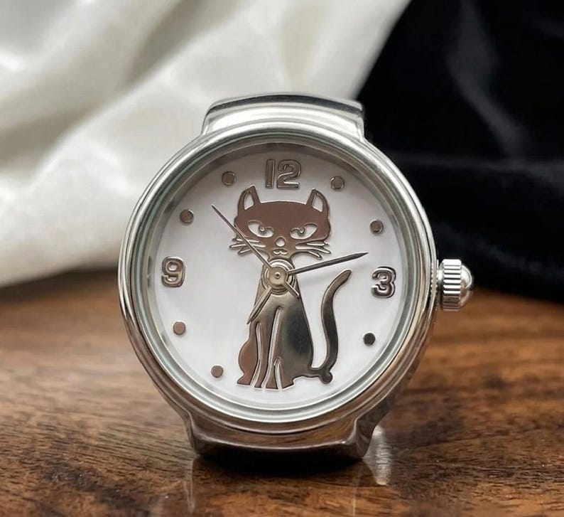 May include: A silver-toned ring watch with a white face. The watch features a cat silhouette at the center, with the numbers 3, 9, and 12 visible. The watch has a small crown on the side and is set on a wooden surface.