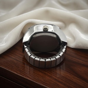 May include: A silver-toned ring watch with a round face and an expandable band. The watch sits on a dark wooden surface with a white fabric in the background. The watch has a small button on top.