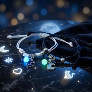 Könnte beinhalten: Zwei verstellbare Kordelarmbänder, eines in Weiß und eines in Schwarz, mit silberfarbenen Metallperlen, einer leuchtenden Kugelperle und einem Mondsichel-Anhänger mit Sternausschnitten in Dunkelblau beziehungsweise Hellblau.
