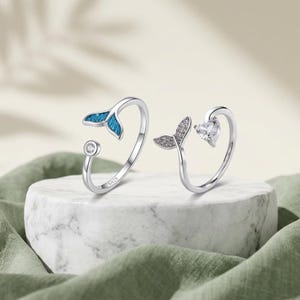 May include: Two open-style silver rings featuring S925 hallmarks. One ring displays a blue enameled mermaid tail and a small round clear stone. The second ring features a pavé-set crystal mermaid tail and a heart-shaped clear stone.