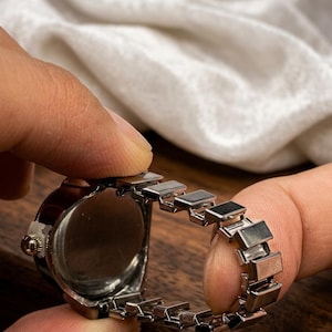 May include: A silver-toned ring watch with a dark brown face and a linked band. The watch is held between two fingers, showcasing the intricate design and the watch's small size. The background is a blurred wooden surface and a white fabric.